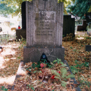 pionowa płyta nagrobna na cokole z tablicą epitafijną i krzyżem łacińskim, obmurowanie mogiły ziemnej pokryte suchymi liśćmi, wokół teren cmentarza