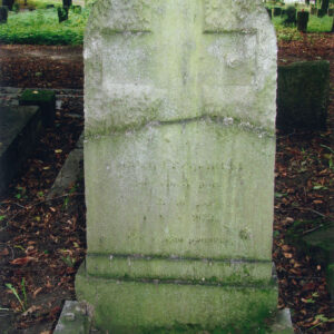 kamienna, omszała tablica z zamazanymi napisami w dolnej części i krzyżem w górnej, porowatej części, z tyłu teren cmentarza, inne nagrobki i drzewa