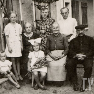 grupa ludzi w różnym wieku, część z nich siedzi, część stoi, pozujących na tle budynku mieszkalnego