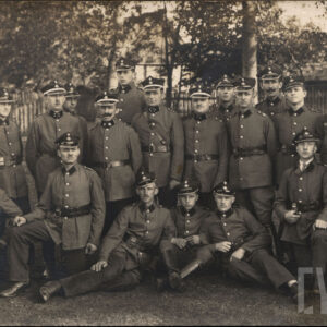 grupa mężczyzn w różnym wieku ubranych w mundury i czapki strażackie, pozujących na tle drewnianego plota i drzew, w głębi widoczne zarysy budynku mieszkalnego 