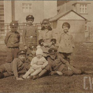 grupa dzieci pozuje do zdjęcia, czwórka siedzi na trawie, pozostała czwórka stoi za nimi, pięciu chłopców ma na głowach równe czapki z emblematem, w głębi fragmenty budynków