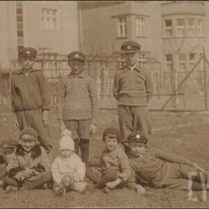 grupa dzieci pozuje do zdjęcia, piątka siedzi na trawie, pozostała trójka stoi za nimi, pięciu chłopców ma na głowach równe czapki z emblematem, w głębi fragmenty budynków mieszkalnych