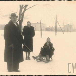 po lewej stoi starszy pan o lasce, obok stoi młoda kobieta, na sankach dwie dziewczynki, w tle drzewa w zimowej szacie i zabudowania