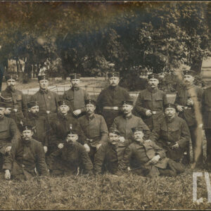 grupa mężczyzn w mundurach polowych, pozująca do fotografii na tle drzew, w głębi widoczne fragmenty zabudowań