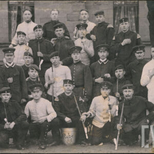 grupa mężczyzn w mundurach polowych pozująca do fotografii na tle ceglanego budynku; część z mężczyzn pozuje trzymają w rękach lub ustach fajki; jeden żołnierz nosi nosi pikielhaubę i pozuje z karabinem na ramieniu 