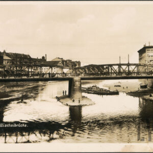 most z żelaznymi przęsłami, pod nim rzeka, z lewej fragment wysokiej kamienicy, za nią niskie zabudowania, z prawej ceglano-drewniany budynek, pod mostem przy środkowym przęśle, na piaszczystej wysepce kilka osób łowi ryby, nieco dalej zacumowane łodzie