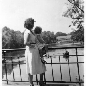 kobieta w sukience stoi przy balustradzie mostku nad stawem, ręką trzyma dziecko siedzące na balustradzie, w głębi drzewa i budynek restauracji