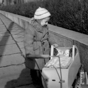 dziewczynka w czapeczce i płaszczyku stoi pochylona nad wózkiem z lalką na chodniku ograniczonym niskim murkiem