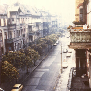 ulica z perspektywy wyższego piętra kamienicy, po lewej stronie rząd kamienic, u ich podnóża szpaler drzew, po prawej fragment balkonu