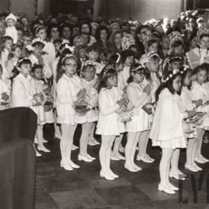 grupa na biało ubranych dziewczynek z koszyczkami z kwiatkami zawieszonymi na wstążkach na szyjach, w białymi wiankami na głowie stoi w rzędach we wnętrzu kościoła