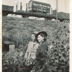 dwie kobiety w kapeluszach stoją w zaroślach, w głębi nasyp kolejowy i przejeżdżający pociąg towarowy