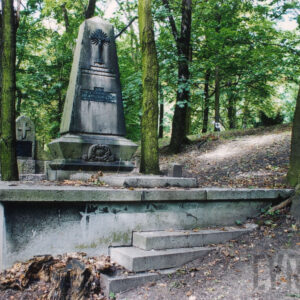 wysoki pomnik w kształcie obelisku umieszczony na kamiennej podstawie, wokół stok cmentarza i zieleń drzew