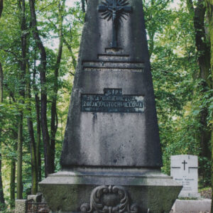 wysoki pomnik nagrobny zwężający się ku górze z napisami i krzyżem łacińskim, za nim zieleń drzew