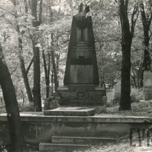 wysoki nagrobek z krucyfiksem ustawiony na cokole, wokół wysokie drzewa i inne nagrobki