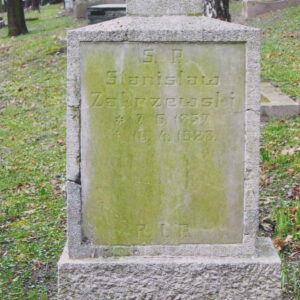 Prostokątna stela z tablica epitafijną pokrytą zielonym nalotem. Wokół inne nagrobki i pnie drzew.