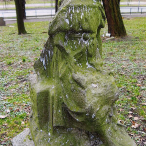 figura anioła pozbawiona głowy i prawej ręki pokryta zielonym nalotem, wokół teren cmentarza, pnie drzew i fragment metalowego ogrodzenia