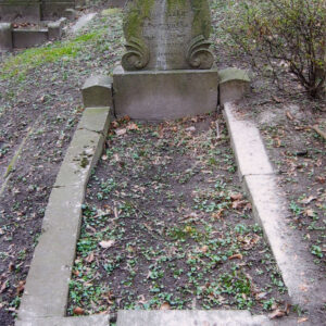 płyta nagrobna pokryta zielonym nalotem i obmurowana mogiła ziemna, wokół teren cmentarza i fragmenty innych nagrobków