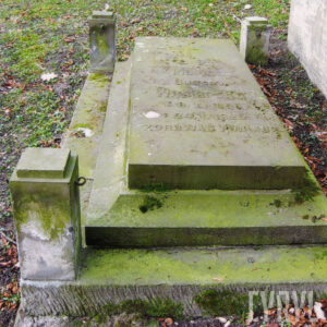 Prostokątna tumba pokryta zielonym nalotem, po bokach czworoboczne słupki. Wokół zielony teren cmentarza.