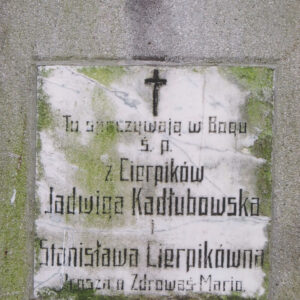 Biała, kwadratowa tablica z czarnymi napisami i krzyżem łacińskim, umieszczona na pionowej płycie nagrobnej. 