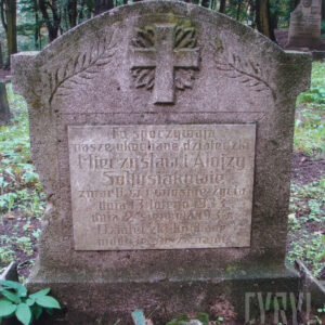 kamienna stela pokryta czarnym i zielonym nalotem, wokół teren cmentarza, fragmenty nagrobków i pnie drzew