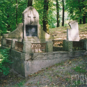 wysoka stela ze spływami wolutowymi po bokach, na niej czarna tablica epitafijna, powyżej której znajduje się krzyż łaciński, wokół teren cmentarza, fragmenty nagrobków i drzewa