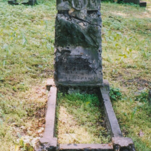 wysoka płyta nagrobna, w którą wtopiony jest krzyż łaciński z płaskorzeźbioną głową Chrystusa, wokół teren cmentarza, inne nagrobki i pnie drzew