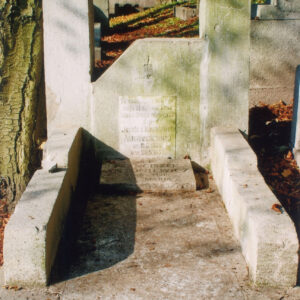 jasnoszary, masywny nagrobek ze stelą o ściętym lewym, górnym narożu i dwoma słupami po bokach, pośrodku tablica epitafijna, z tyłu teren cmentarza