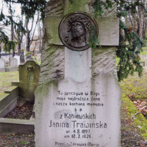 pionowa stela ciosana w górnej części, na niej wtopiony krzyż z płaskorzeźbą Chrystusa i tablicą epitafijną, z tyłu fragmenty innych nagrobków i drzewa