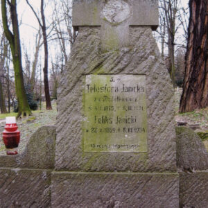 wysoka, ciosana stela z wtopionym krzyżem łacińskim i śladem po tondzie, w dolnej części tablica epitafijna, na krawędzi płyty stoi czerwony znicz, z tyłu drzewa