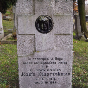 prostokątna pionowa płyta nagrobna z płaskorzeźbionym krzyżem i napisami, na przecięciu ramion krzyża okrągły medalion z wyobrażeniem głowy Chrystusa w koronie cierniowej, za płytą fragmenty nagrobków i pnie drzew