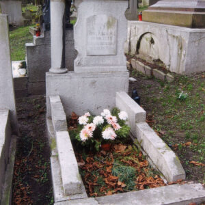 obmurowana mogiła, na zamknięciu zagłówka wysoki cokół, na nim stela złożona z prostokątnej płyty i odsuniętej od niej kolumny podpierających tympanon zwieńczony spłaszczonym łukiem, za nagrobkiem fragmenty kamiennych grobów, w głębi drzewa