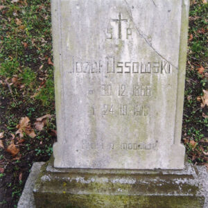 prostokątna kamienna tablica z białego marmuru z wyrytym napisem, pęknięta w górnym prawym narożniku, ustawiona lekko pochyło na niskim cokole