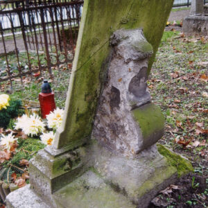 tył prostokątnej tablicy ustawionej lekko pochyło na konsoli z piaskowca z licznymi ubytkami i zielonym nalotem, przed tablicą kwiaty i znicz, w głębi fragment metalowego ogrodzenia