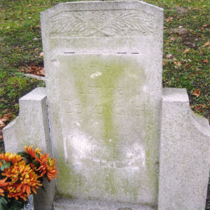 prostokątna płyta ze ściętymi narożnikami ustawiona na cokole, po jej bokach niższe słupki o ściętych wklęsło krawędziach bocznych, w górnej części płyty płaskorzeźba przedstawiająca dwie krzyżujące się gałązki palmowe, na płycie zielony nalot, z lewej fragment kamiennego wazonu z kwiatami, za nagrobkiem trawa