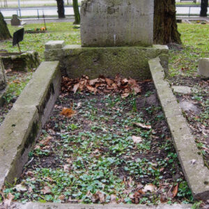 ziemna mogiła obmurowana, w części zagłówkowej tablica pionowa o ściętej ukośnie górnej krawędzi z odłamanym lewym narożnikiem i słabo czytelnym napisem, całość pokryta zielonym nalotem, za nagrobkiem trawa, w głębi pnie drzew