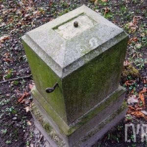 czworoboczny kamienny postument widziany od góry, na jego szczycie, pośrodku metalowy fragment osadzony w kamieniu, prawdopodobnie pozostałość po nieistniejącym krzyżu, z lewej strony metalowy uchwyt dawnego ogrodzenia, postument osadzony na uskokowo rozszerzającym się cokole, całość pokryta zielonym nalotem