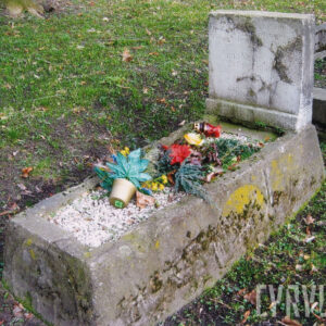 prostokątna mogiła ziemna, obwiedziona płaskorzeźbionym, wysokim kamiennym obmurowaniem w typie sarkofagu zwężającego się ku górze, bez wieka, środek wypełniony białymi drobnymi kamykami i pozostałościami sztucznych wiązanek, w części zagłówkowej prosta, pionowa płyta z wyrytym słabo czytelnym napisem, całość mocno zniszczona, z licznymi ubytkami i pokryta mchem