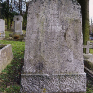 trapezoidalna, lekko zwężająca się ku górze tablica nagrobna zwieńczona spłaszczonym z dwoma uskokami, ustawiona na wysokim, dwustopniowym cokole, na tablicy ryta inskrypcja, częściowo zatarta, w głębi murowane nagrobki i pnie drzew