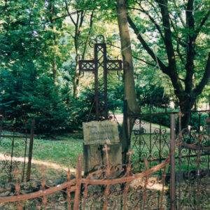 mogiła ziemna ogrodzona ozdobną żeliwną kutą kratą, pośrodku, metalowy, ażurowy krzyż łaciński z trójlistnie zakończonymi ramionami, pod nim lekko odchylona do tyłu tablica inskrypcyjna, w głębi drzewa