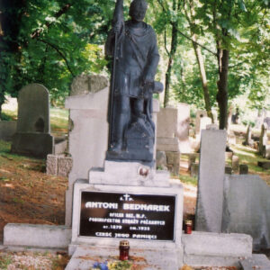 obmurowany nagrobek z kamienną płytą pośrodku, po bokach wysypane białe kamyki, między nimi chwasty, na cokole z czarną tablicą epitafijną figura św. Floriana, biały napis na tablicy