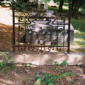 fragment kutego ogrodzenia w formie prostych i zakręconych spiralnie linii umieszczonym na podmurówce, za nim dość wysoki prostokątny murowany nagrobek w formie prostopadłościanu z widocznymi cegłami, na nim pochylona do tyłu tablica, całość widziana od tyłu