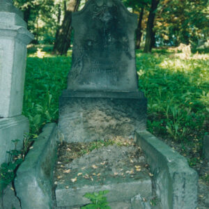 zniszczony kamienny nagrobek otoczony wysokim obramowaniem, z lewej przechylonym, w części zagłówkowej stela zakończona łukiem wklęsło-wypukłym