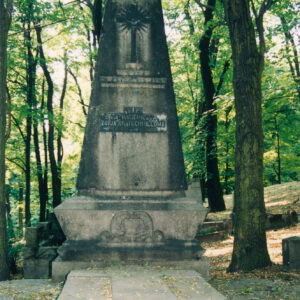 nagrobek przykryty kamienną płytą z płaskorzeźbionym krzyżem łacińskim, w części zagłówkowej monumentalny czworoboczny słup zwężający się ku górze, posadowiony na sarkofagowej podstawie na nim płaskorzeźbiony krzyż z glorią, poniżej inskrypcja, w głębi drzewa