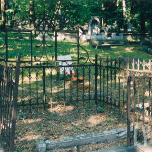 mogiła ziemna okolona żeliwną ozdobną kratą osadzoną na niskiej podmurówce, część środkowa podwyższona z fantazyjnie zakręconymi liniami i motywem roślinnym, brak tablic inskrypcyjnych, w głębi kamienne nagrobki i drzewa