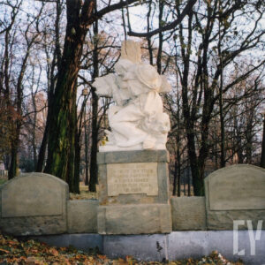 nagrobek w formie tryptyku, pośrodku rzeźba mężczyzny w długiej szacie, z kapeluszem na plecach obejmującego krzyż stojący na postumencie