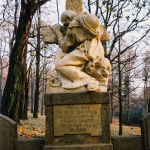 pomnik mężczyzny w długiej szacie, z kapeluszem na plecach obejmującego krzyż stojący na postumencie, poniżej tablica inskrypcyjna