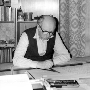 mężczyzna w białe koszuli i ciemnej kamizelce siedzi w pokoju przy desce kreślarskiej, za plecami mężczyzny segment z otwartymi i zamkniętymi półkami