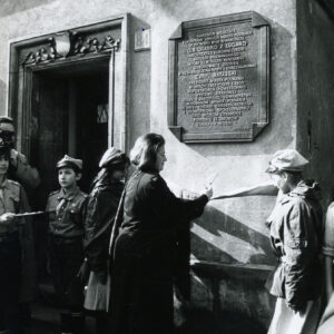 ściana kamienicy z ozdobnym obramieniem drzwi wejściowych i zawieszoną po ich prawej stronie masywną tablicą, przed nią grupa ludzi, w tym harcerze i kobieta przecinająca wstęgę