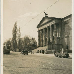 na pierwszym planie brukowana ulica z torami tramwajowymi, jadą po niej dwa samochody i tramwaj, przy ulicy neoklasycystyczny budynek z sześciokolumnowym portykiem, zwieńczony rzeźbą konia ze skrzydłami do którego prowadzą monumentalne schody z rzeźbami po obu stronach