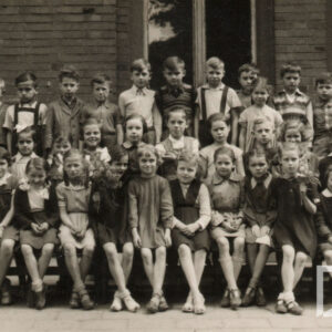 kilkudziesięciu uczniów w trzech rzędach, pierwszy rząd, same dziewczynki, siedzi, pozostałe dwa rzędy stoją, w ostatnim, górnym rzędzie stoją dwie dziewczynki i kilkunastu chłopców, po prawej stronie stoi nauczycielka, w głębi fragment ceglanego muru z oknami